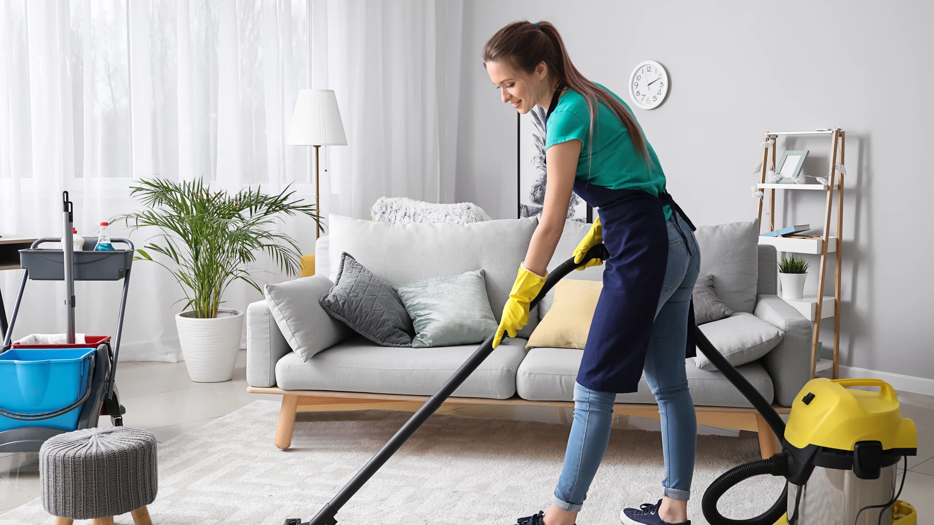 Femme en tenue de nettoyage utilisant un aspirateur jaune sur un canapé gris dans un salon moderne et lumineux